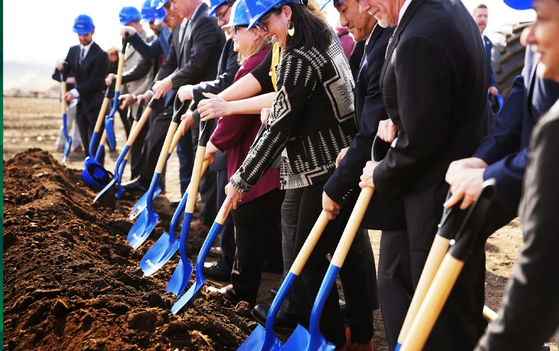 People in blue hard hats using shovels at a groundbreaking ceremony.